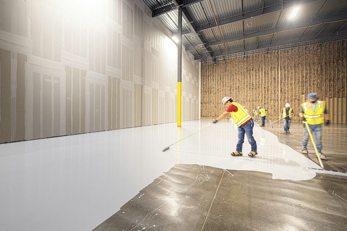 Crew in safety vests spreading a wet white epoxy coating across a warehouse slab with squeegees. Reference photo of coating in progress.